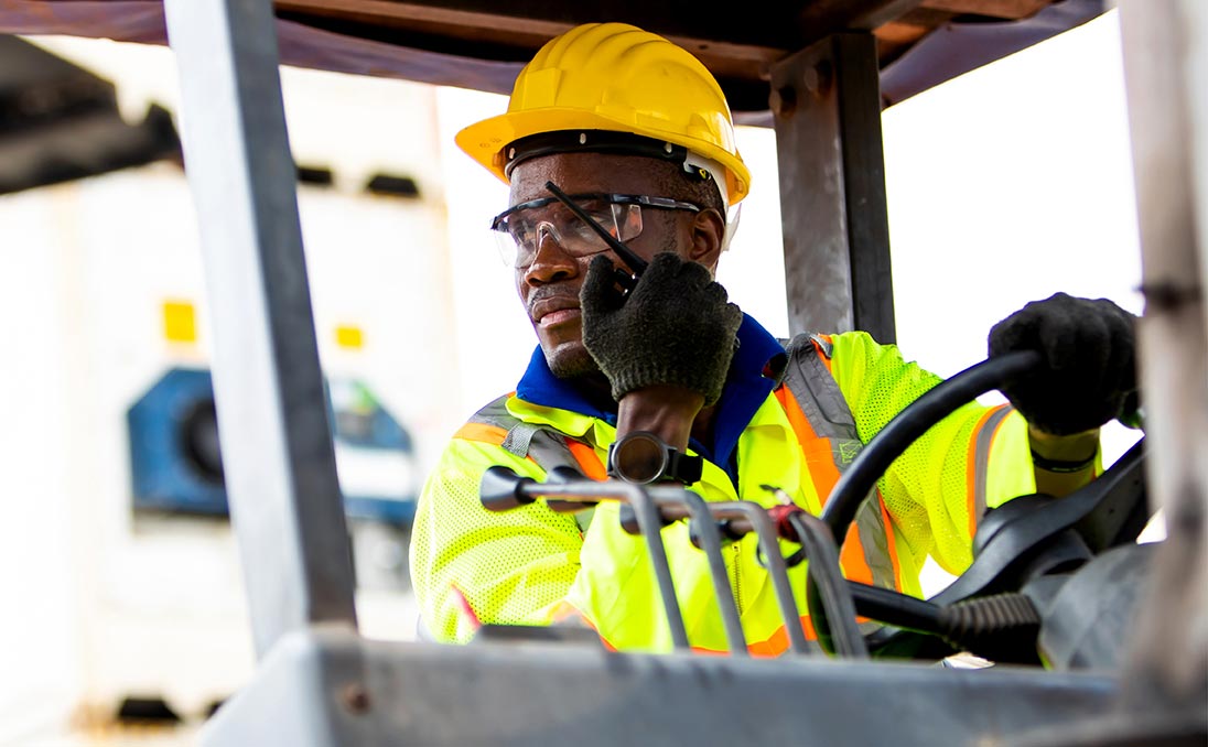 A man driving a fork lift on a walkie-talkie wearing PPE including gloves goggles and a hard hat