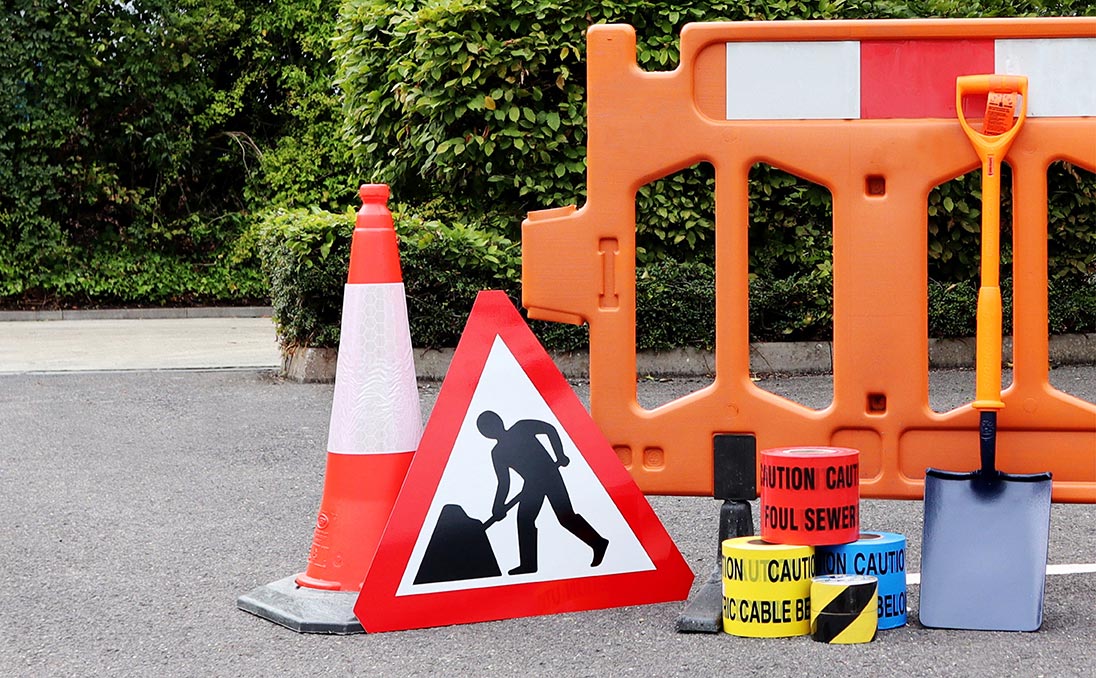 An orange barrier fence with a traffic cone and traffic signs, including caution tape and a spade