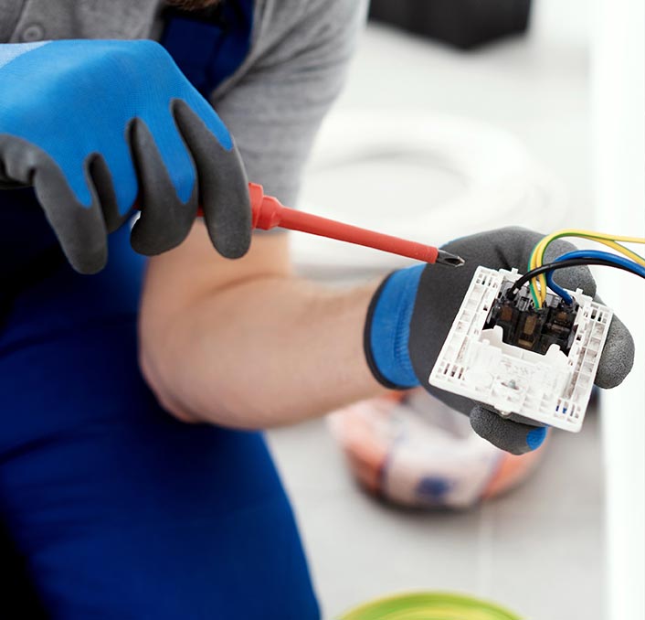 Close up on a hand in gloves holding a screwdriver in one hand and an electrical outlet in his other