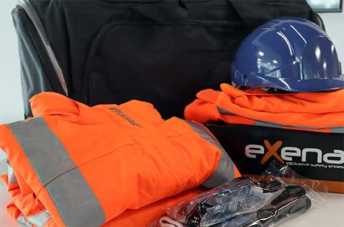 A black bag is behind orange hivis jackets, a shoe box, hard hat, gloves and goggles in plastic wrappers.